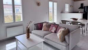 A modern living room with a grey sofa, white coffee table, and a vase of dried flowers. A kitchen is visible in the background with a black range hood. - The Oceanid (Saint-Denis-d'Oléron)