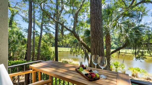 Un tavolo in legno con un vassoio di frutta e bicchieri da vino, con vista su uno stagno circondato da alberi.