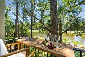 Una mesa de madera con una bandeja de frutas y copas de vino, con vista a un estanque rodeado de árboles.