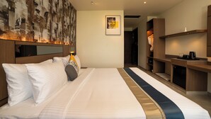 Hotellihuone, jossa on suuri sänky, puinen pääty, seinälle kiinnitetty taideteos, kehystetty kuva ja pieni keittotila.