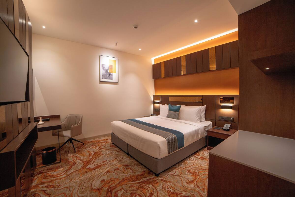 A hotel room with a bed, bedside table, chair, desk, and a painting on the wall.