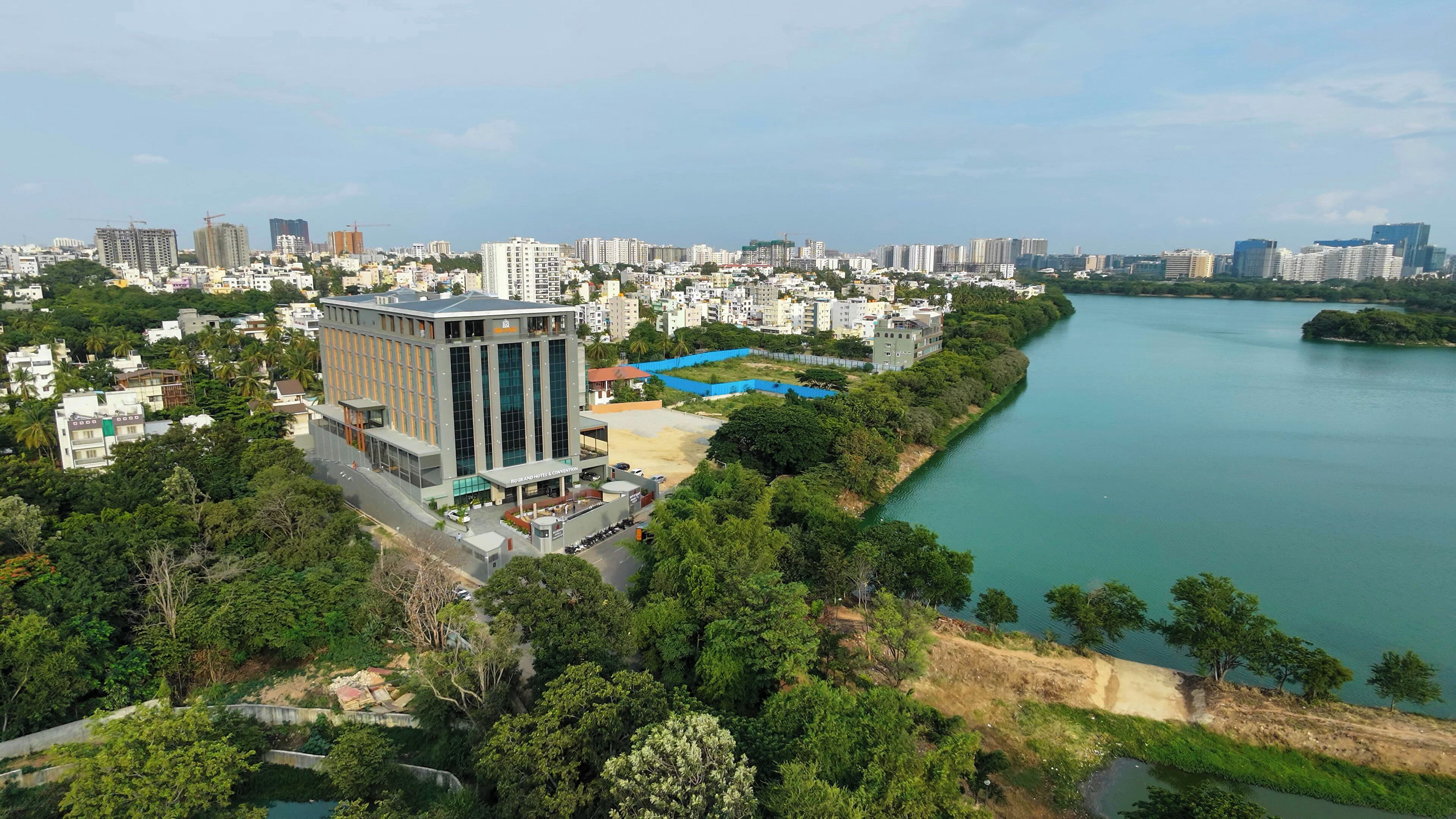 A modern hotel with a glass facade, situated beside a river, surrounded by greenery and urban buildings.