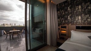 Hotellihuone, jossa on suuri liukuovi, joka johtaa ulkotilaan, jossa on istuinalue. Huoneesta on näkymä kaupunkiin, ja siinä on valkoisilla tekstiileillä varustettu sänky sekä kaupunkiaiheinen tapetti.