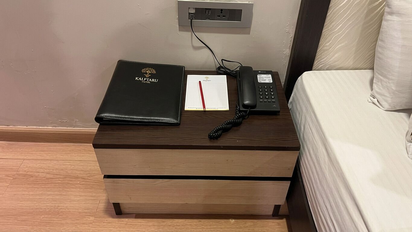 Ein Hotelzimmer mit Nachttisch aus Holz, einer Kalptaru Hotel Mappe, einem Notizblock mit rotem Stift, einem schwarzen Telefon und einem Bett mit weißer Matratze.