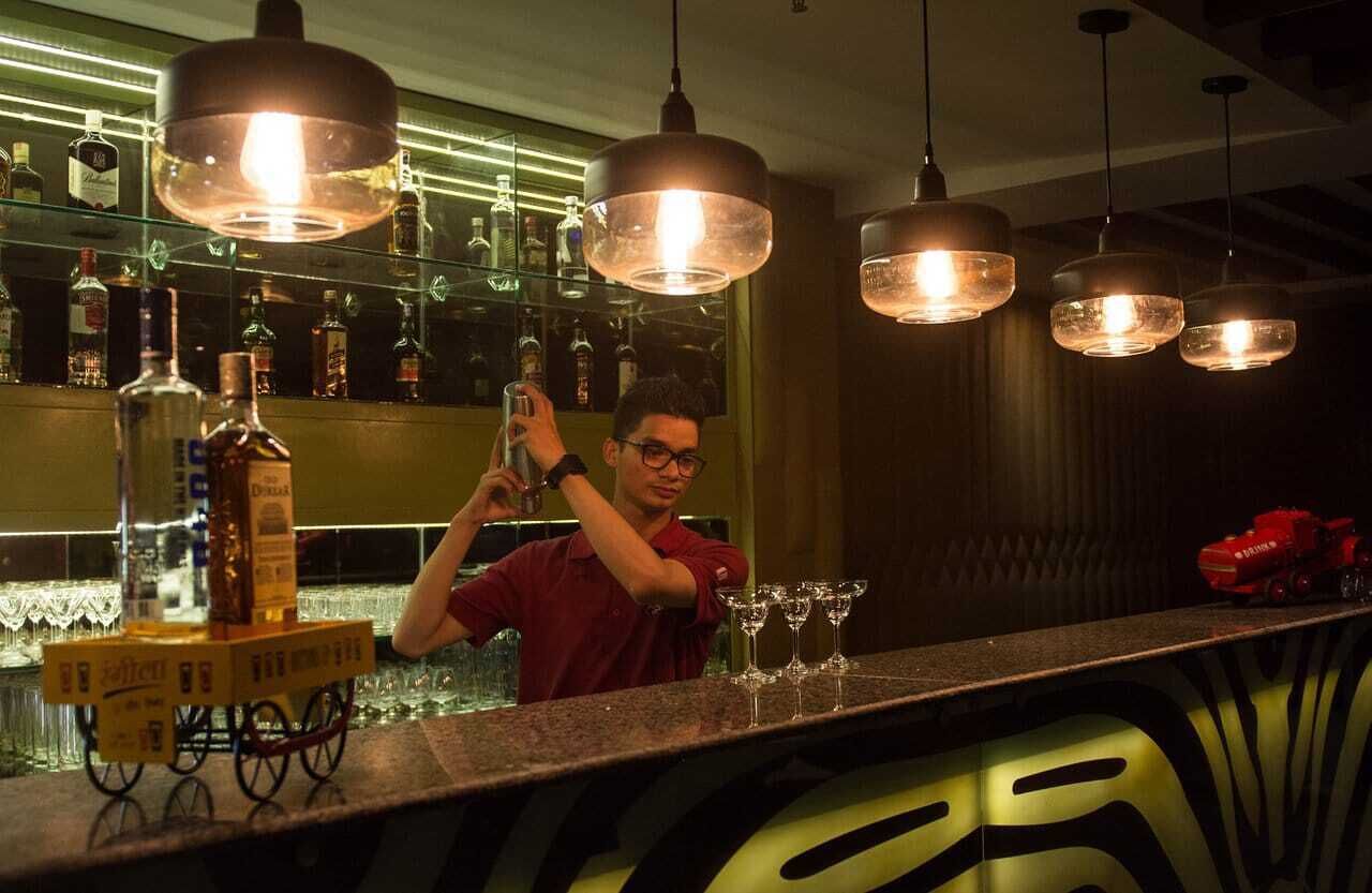 A bar area with hanging pendant lights, a bartender preparing a drink, and a shelf with various bottles.
