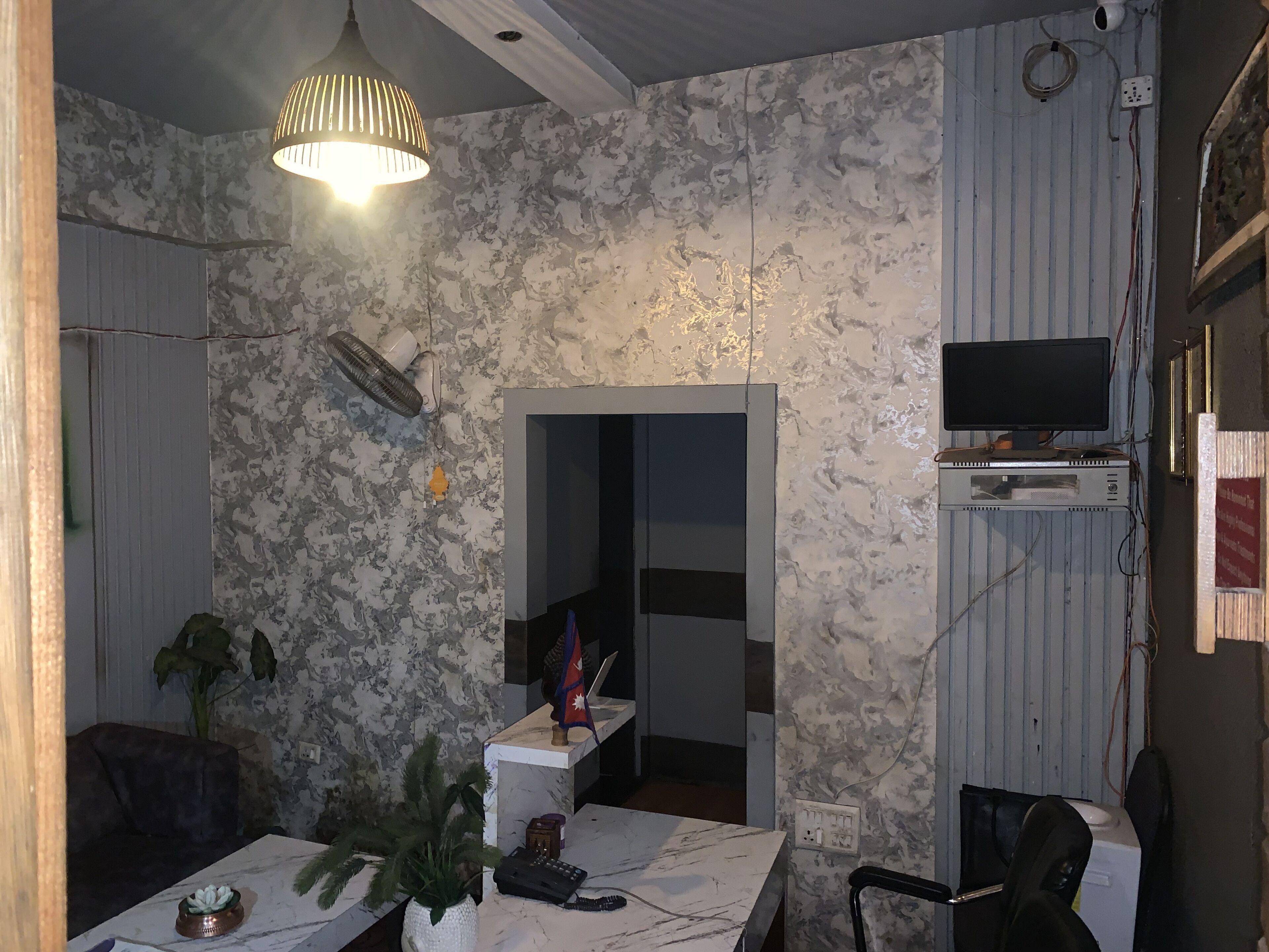 A room with a marble-patterned wall, a ceiling fan, a table with a potted plant, a TV mounted on the wall, and a power outlet.
