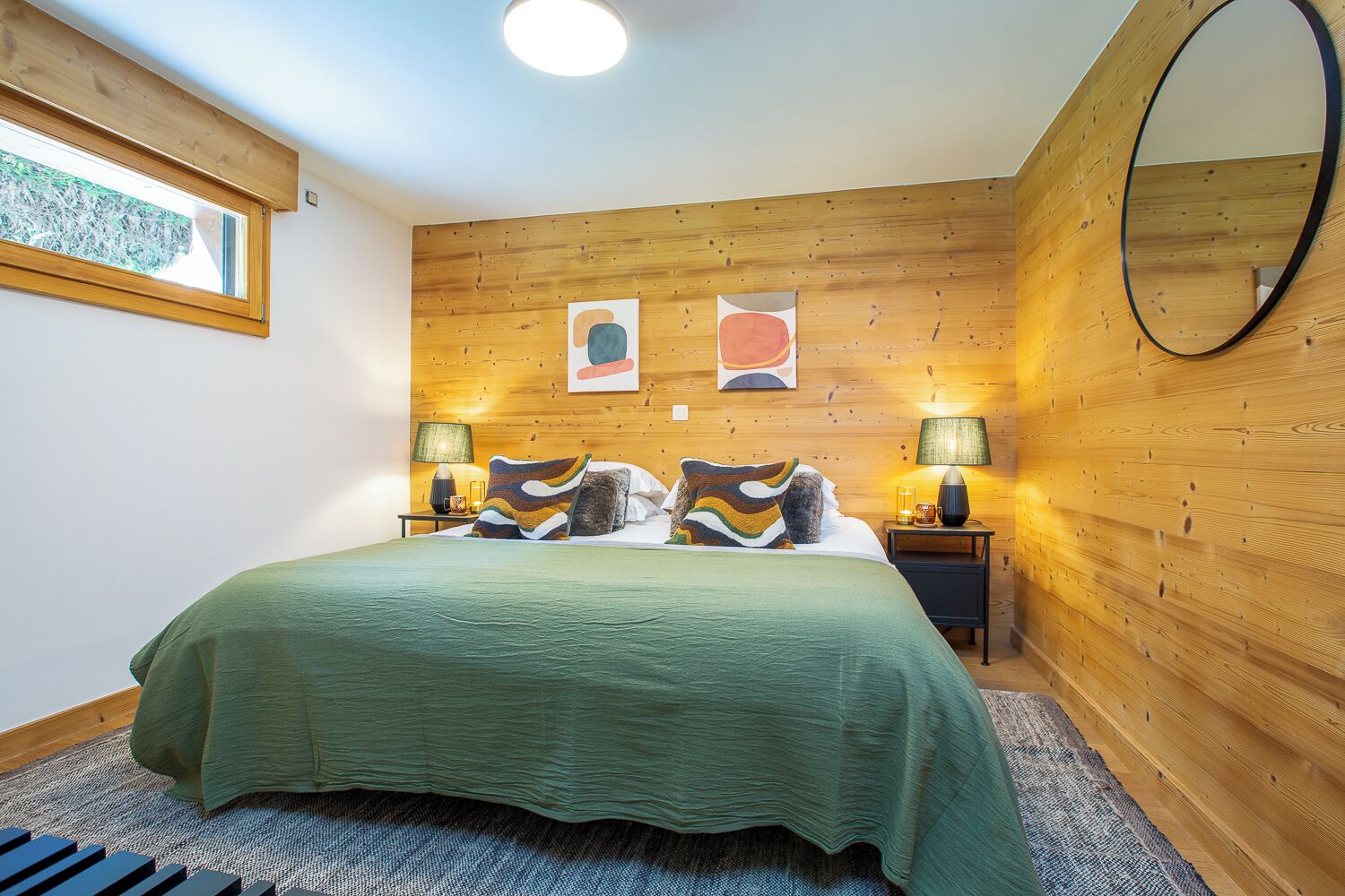 A bedroom with a wooden wall panel, a bed with a green bedspread, two bedside lamps, and a round mirror on the wall.