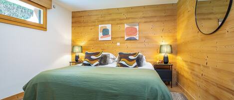 A bedroom with a wooden wall panel, a bed with a green bedspread, two bedside lamps, and a round mirror on the wall.