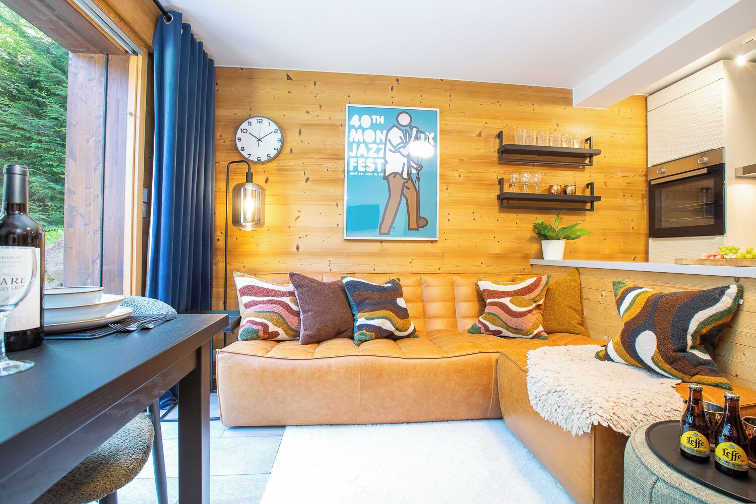 A cozy room with a wooden wall, a sofa with colorful cushions, a clock, and a poster advertising a jazz festival.