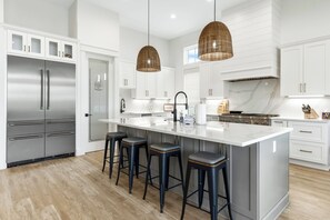 A modern kitchen with white cabinetry, stainless steel appliances, a large island with bar stools, and a hanging wicker light fixture. - 5 BR NEW 2026 BUILD | 5 O'Clock Shadow | Gulf Views | Sleeps 12 (Inlet Beach)