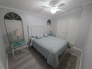 Ein Schlafzimmer mit Bett, Deckenventilator, Nachttisch mit Lampe, Kommode und Spiegel.