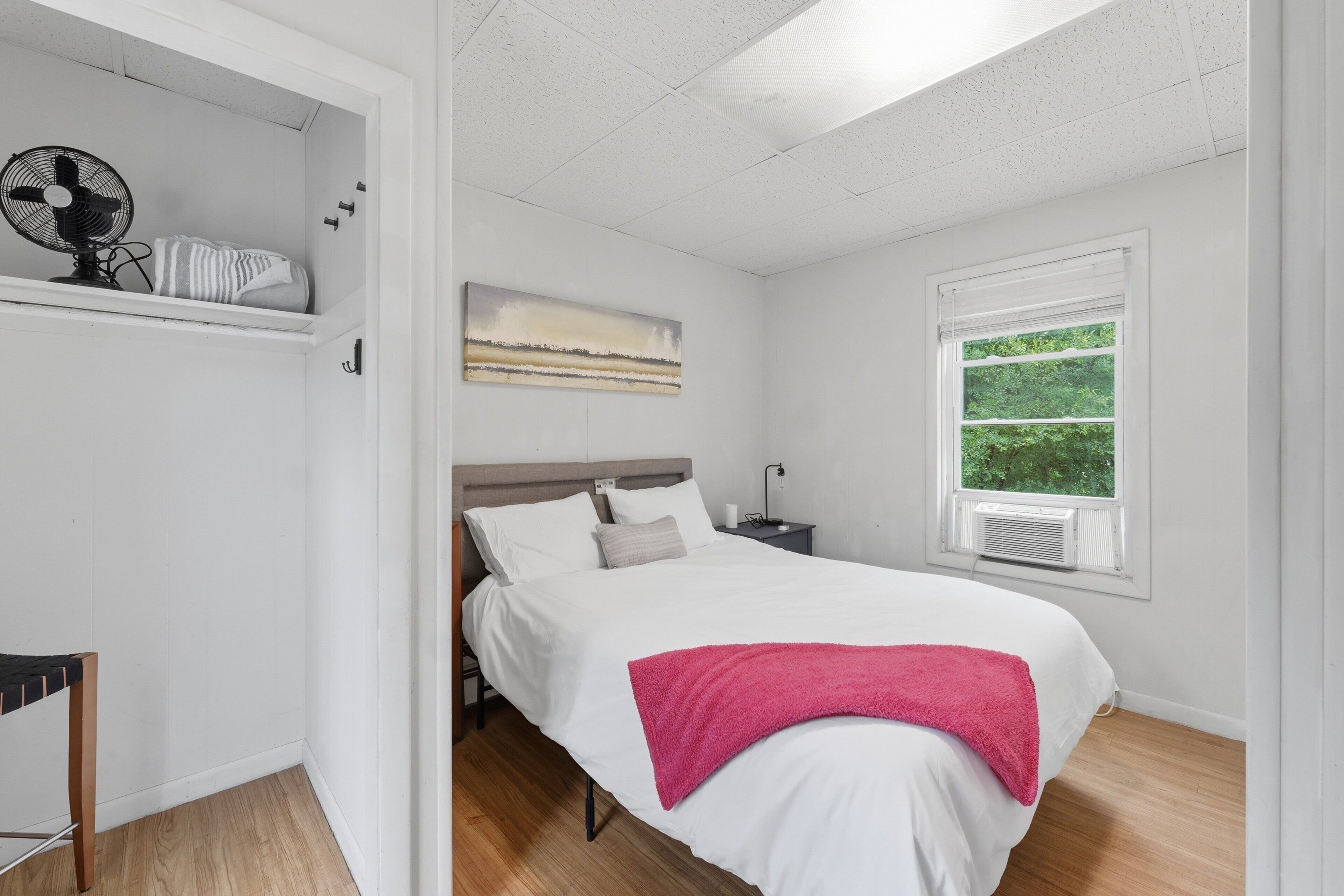 A bedroom with a bed, a nightstand, a wall-mounted fan, a window with a view, and a shelf with a fan and a striped item.