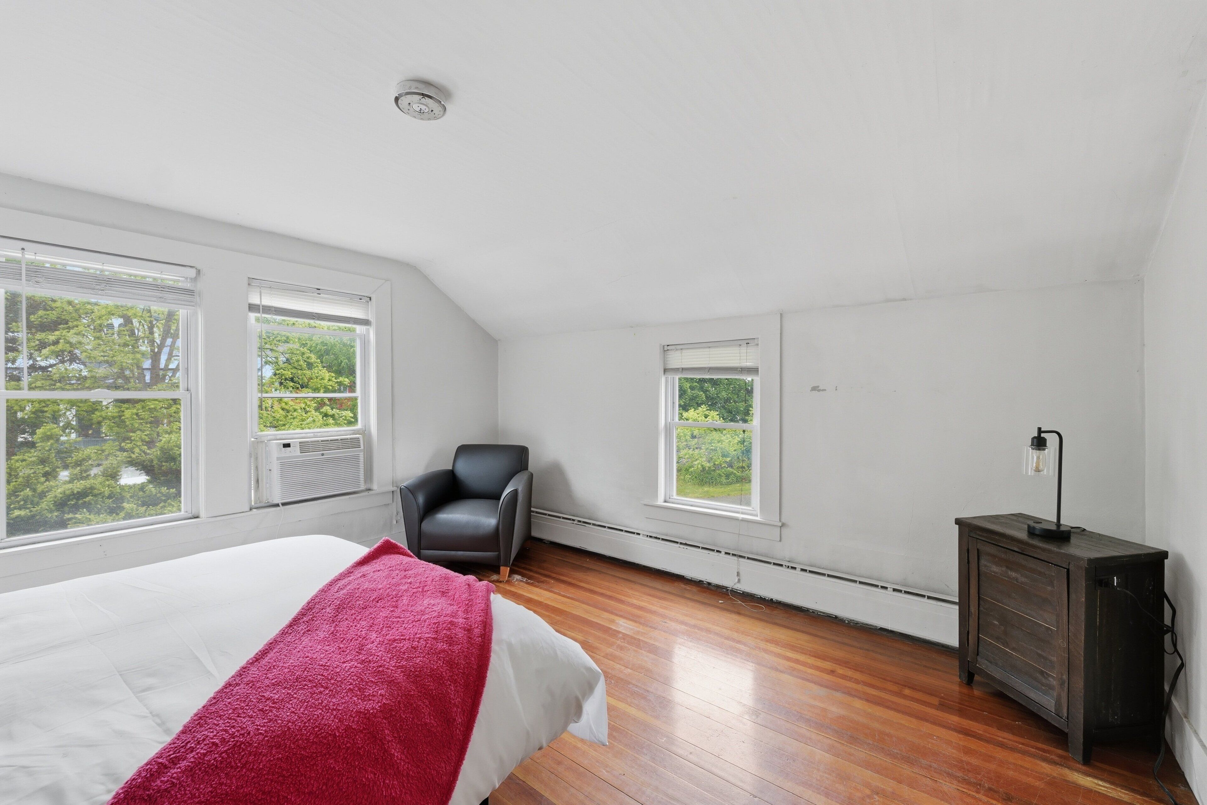 A bedroom with a bed, a chair, a dresser, and large windows with a view of greenery.