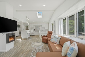 A modern living room with a fireplace, a sofa with decorative pillows, a television, and a kitchen in the background. - Modern Hampton Bays Cottage Near Beaches & Town 2 Bedroom Cottage (Hampton Bays)