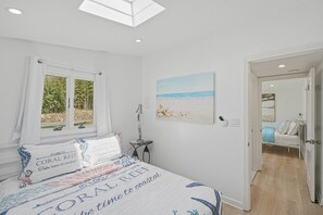 A bedroom with a bed, a window with curtains, a bedside table, and a painting of a beach scene.