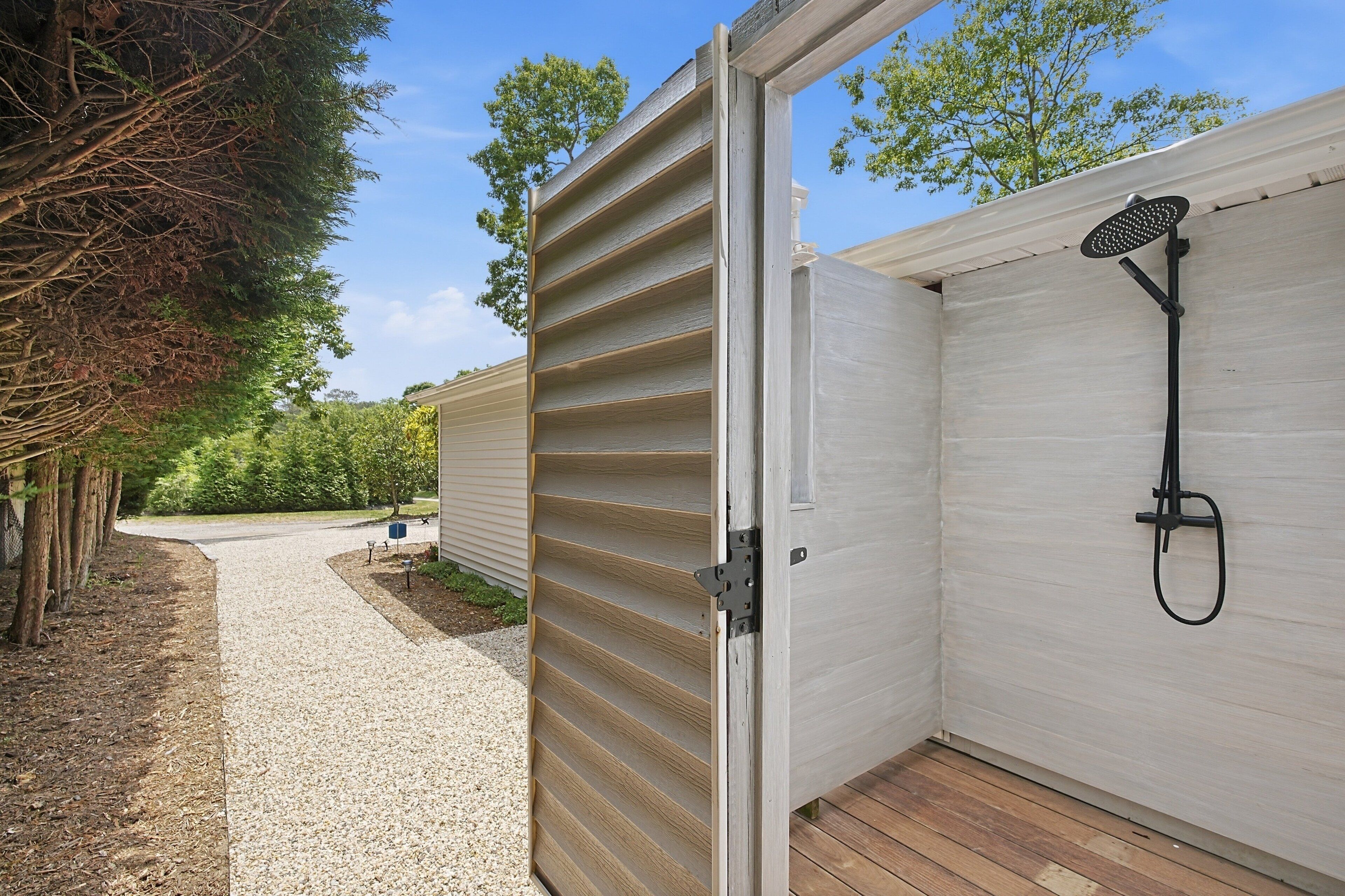 Puerta abierta a un espacio exterior cubierto con ducha, camino de grava y terraza de madera.