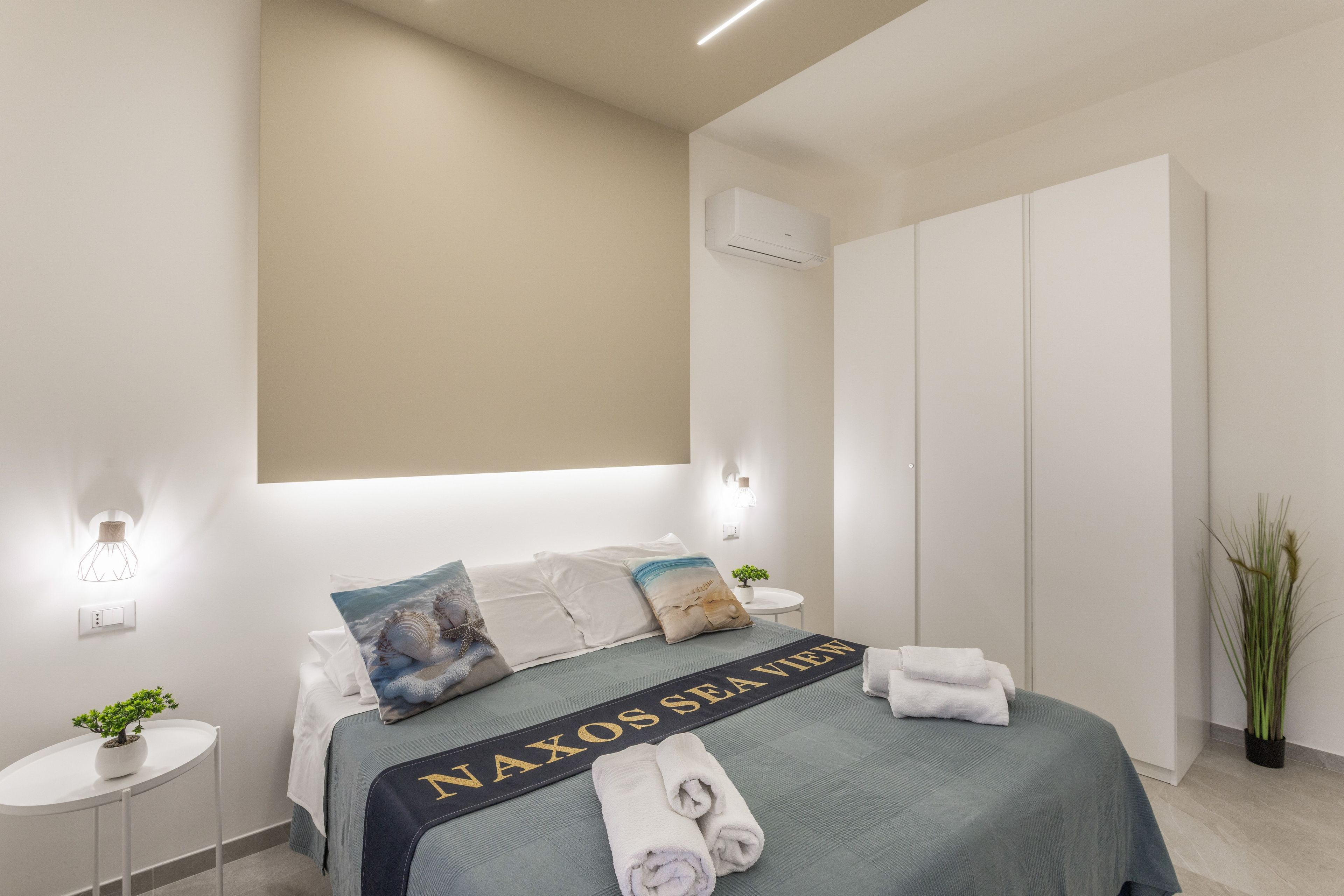 A hotel room with a bed, a bedside table with a plant, a wall-mounted air conditioner, and a large wardrobe.