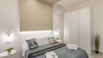 A hotel room with a bed, a bedside table with a plant, a wall-mounted air conditioner, and a large wardrobe.