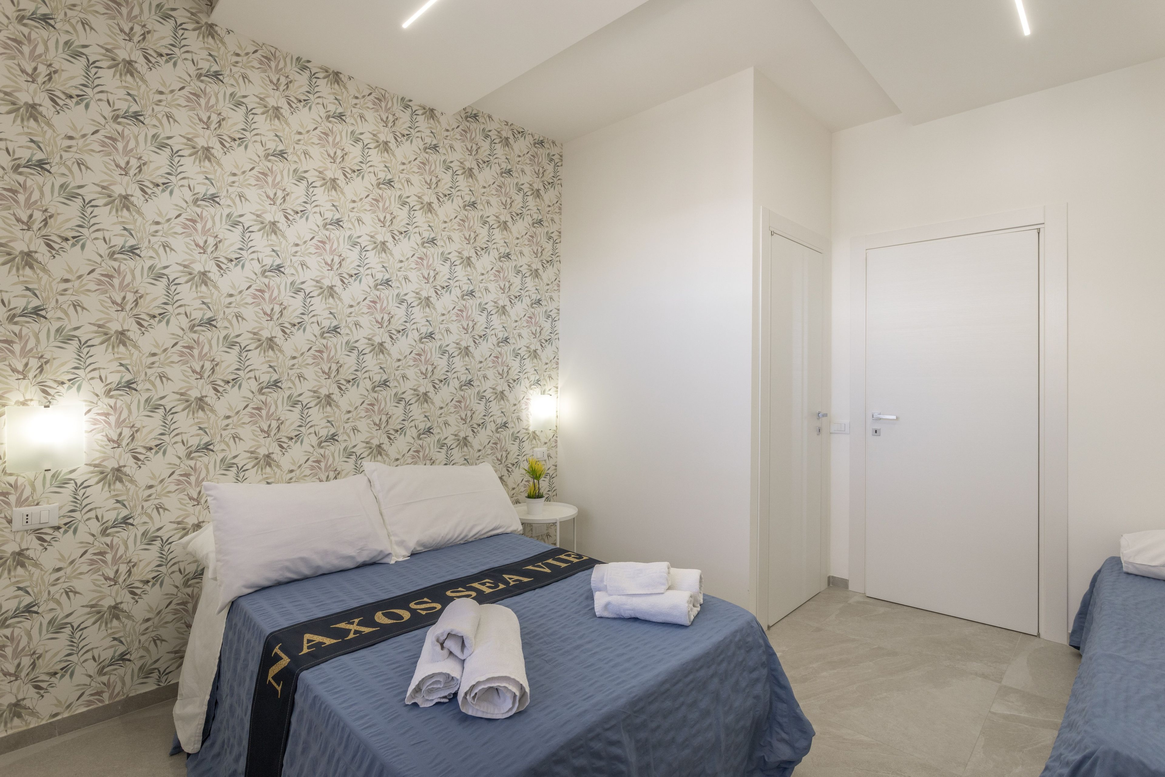 A hotel room with a bed, a nightstand, a wall-mounted light, and a door.