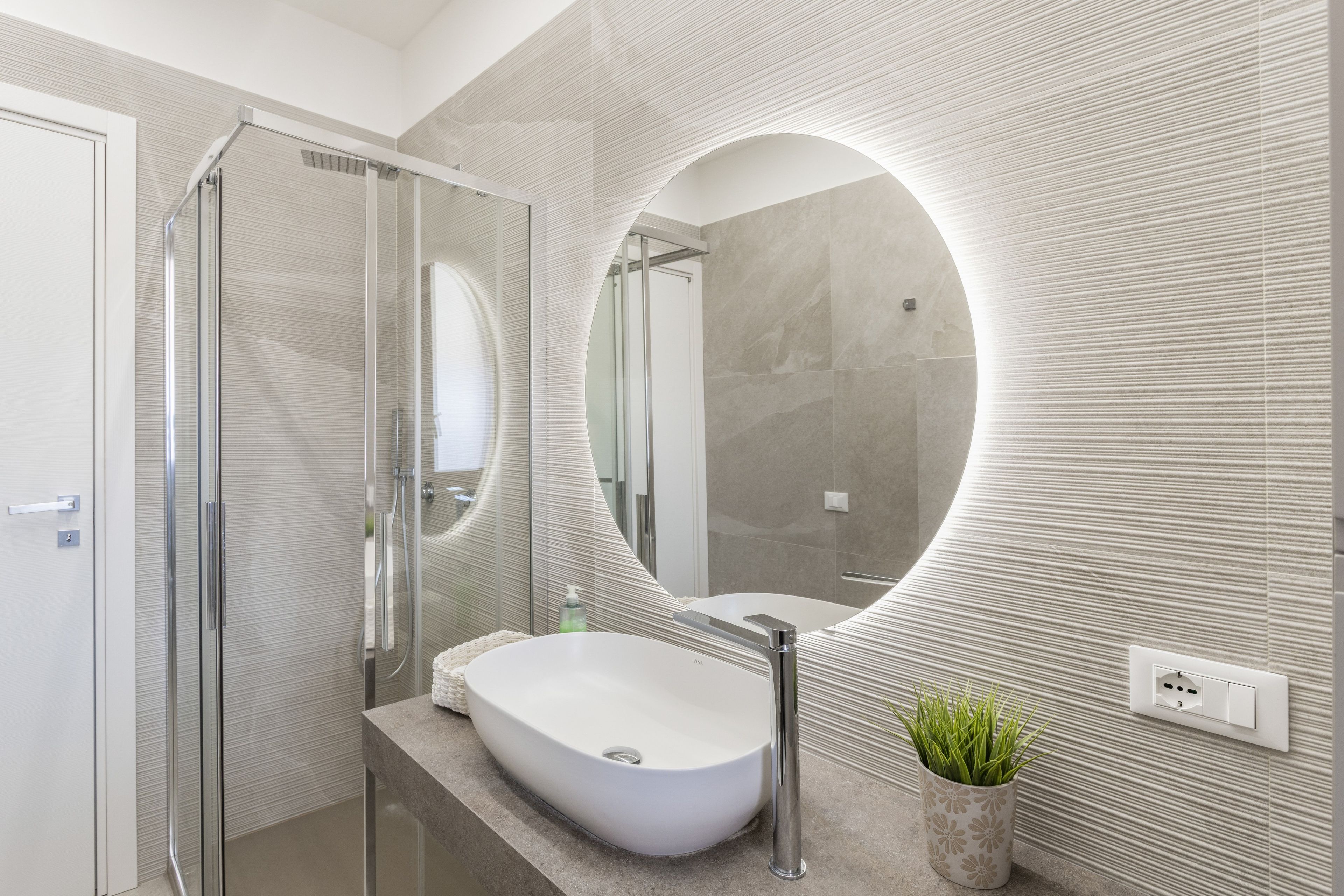 A modern bathroom with a glass shower enclosure, a white sink, and a round mirror with integrated lighting.