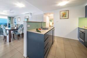 A modern kitchen with a central island, a dining area with chairs, and a view to the outside. - Azure Sea-Renity: Ocean Views, Pool & Gym (Airlie Beach)