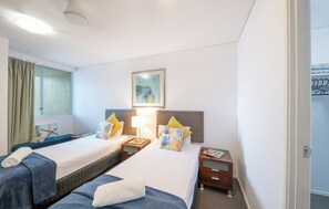 A hotel room with two beds, a window with curtains, a framed picture on the wall, and a nightstand with a lamp. - Azure Sea-Renity: Ocean Views, Pool & Gym (Airlie Beach)