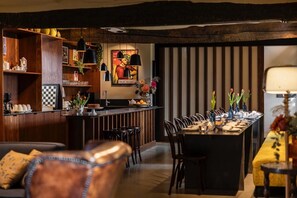 A dining area with a long table, chairs, a bar area with shelves, and a painting on the wall.