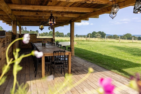 A wooden deck with a long table and chairs, surrounded by a wooden fence and a grassy field.