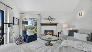 A living room with a fireplace, a sofa, a blue armchair, a round coffee table, and a view of the outdoors. - 4084 Shibui by the Bay - Skyline Forest Townhouse (Monterey)