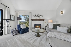 A living room with a fireplace, a sofa, a blue armchair, a round coffee table, and a view of the outdoors. - 4084 Shibui by the Bay - Skyline Forest Townhouse (Monterey)