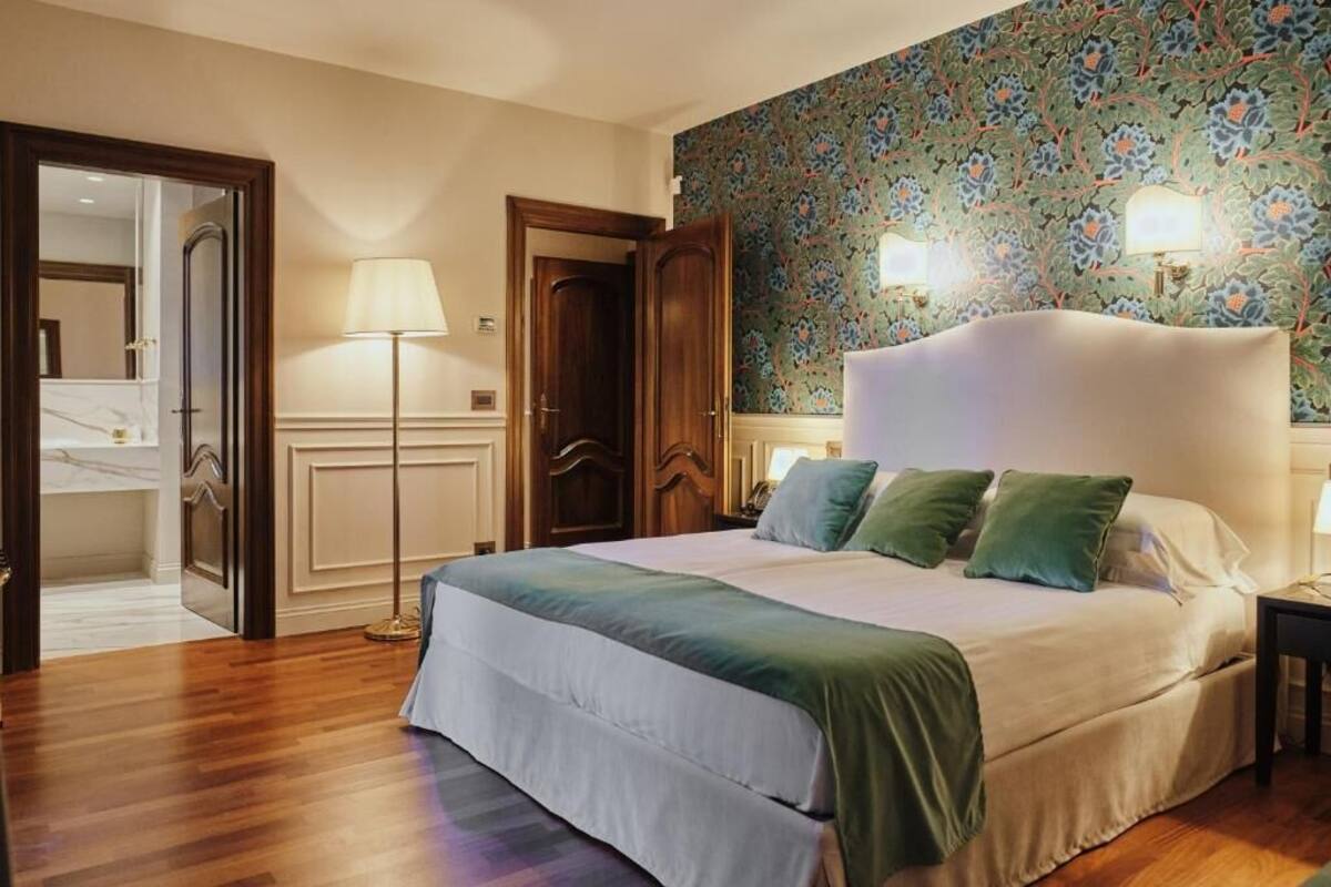 Habitació d'hotel amb un llit gran, un sòl de fusta, una porta que dona a un bany i un fons de pantalla amb motius florals.