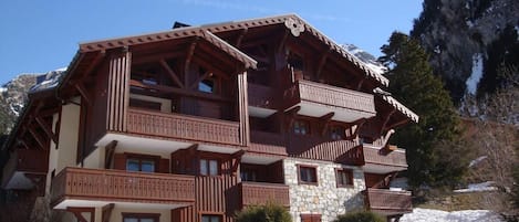 A multi-story chalet with wooden balconies and stone walls, surrounded by snow and mountains.
