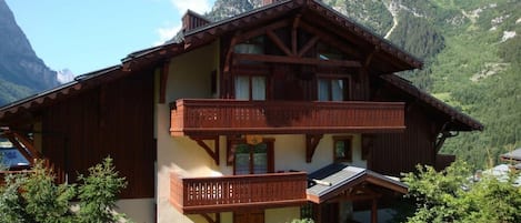 A two-story wooden chalet with balconies, set against a mountainous backdrop.