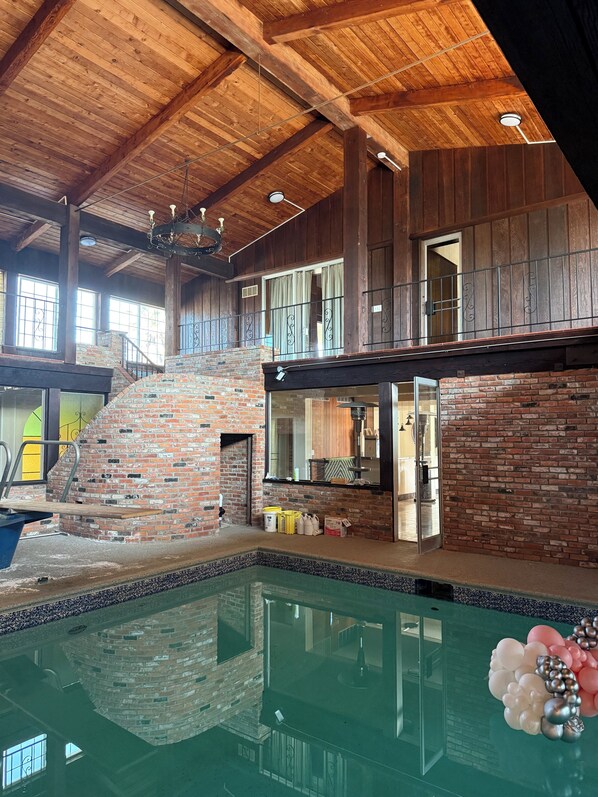 A spacious indoor pool area with a brick fireplace, wooden ceiling beams, and a balcony with metal railings.