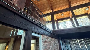 Una habitación con techos altos y vigas de madera a la vista, un balcón con barandillas metálicas y una pared de ladrillo.