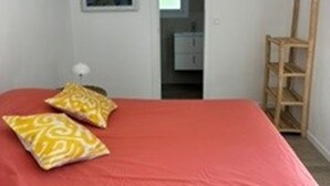 Une chambre avec une couvre-lit rouge, deux coussins à motifs, une échelle en bois, un mur blanc et un tableau encadré.