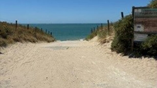 Un sentier de sable qui longe la plage et mène à l’océan, avec un panneau indicateur visible.