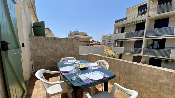 Terraço no telhado com mesa posta para dois, rodeado por cadeiras de plástico branco, e vista para edifícios residenciais ao fundo.