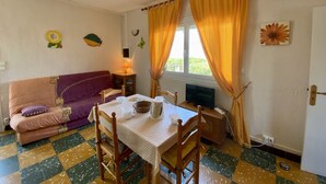 Um quarto acolhedor com um sofá, uma mesa com cadeiras, uma televisão e uma janela com cortinas.