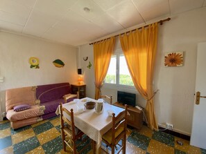 Ein gemütliches Zimmer mit einer Couch, einem Tisch mit Stühlen, einem Fernseher und einem Fenster mit Vorhängen.