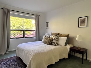 Une chambre à coucher avec un lit, une table de chevet, une lampe, une fenêtre avec des rideaux et un tableau encadré au mur.