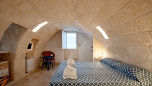 Ein Zimmer mit Steindecke, einem Bett mit gemusterten Textilien, einem Sessel, einem kleinen Schreibtisch und einem Fenster.