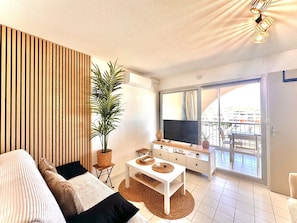 A modern living room with a sofa, a TV, a dining table, a potted plant, and a balcony with a view. - Cozy apartment, AC, canal view (Sète)