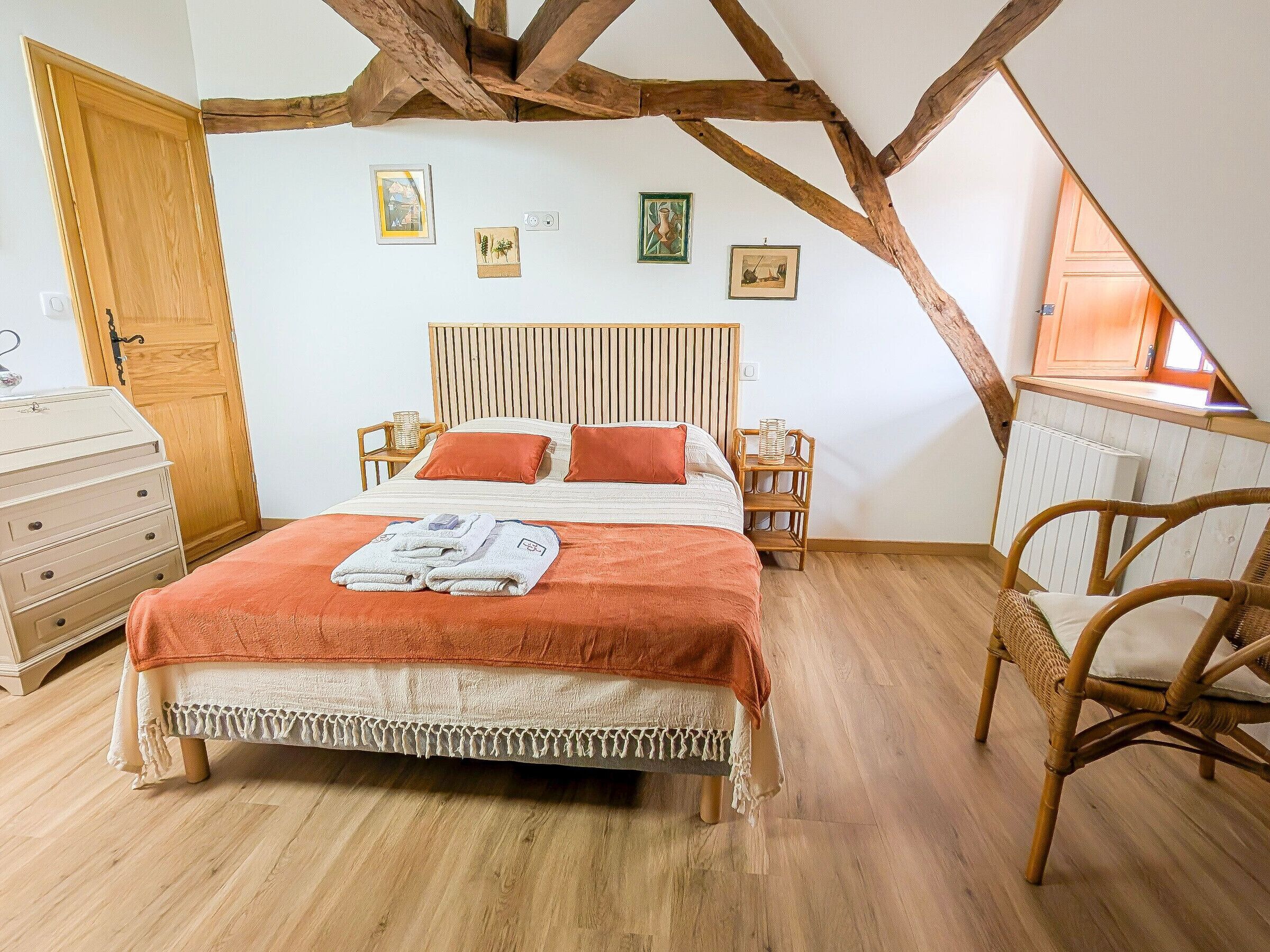 Une chambre avec un plafond en bois, un lit recouvert d’une courtepointe blanche et orange, une commode en bois et une chaise en osier.