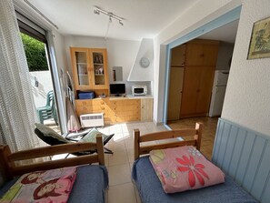 A room with a bed, a small desk, a chair, a kitchenette, and a door leading to another room. - Cozy apartment with pool - 3 guests (Argelès-sur-Mer)