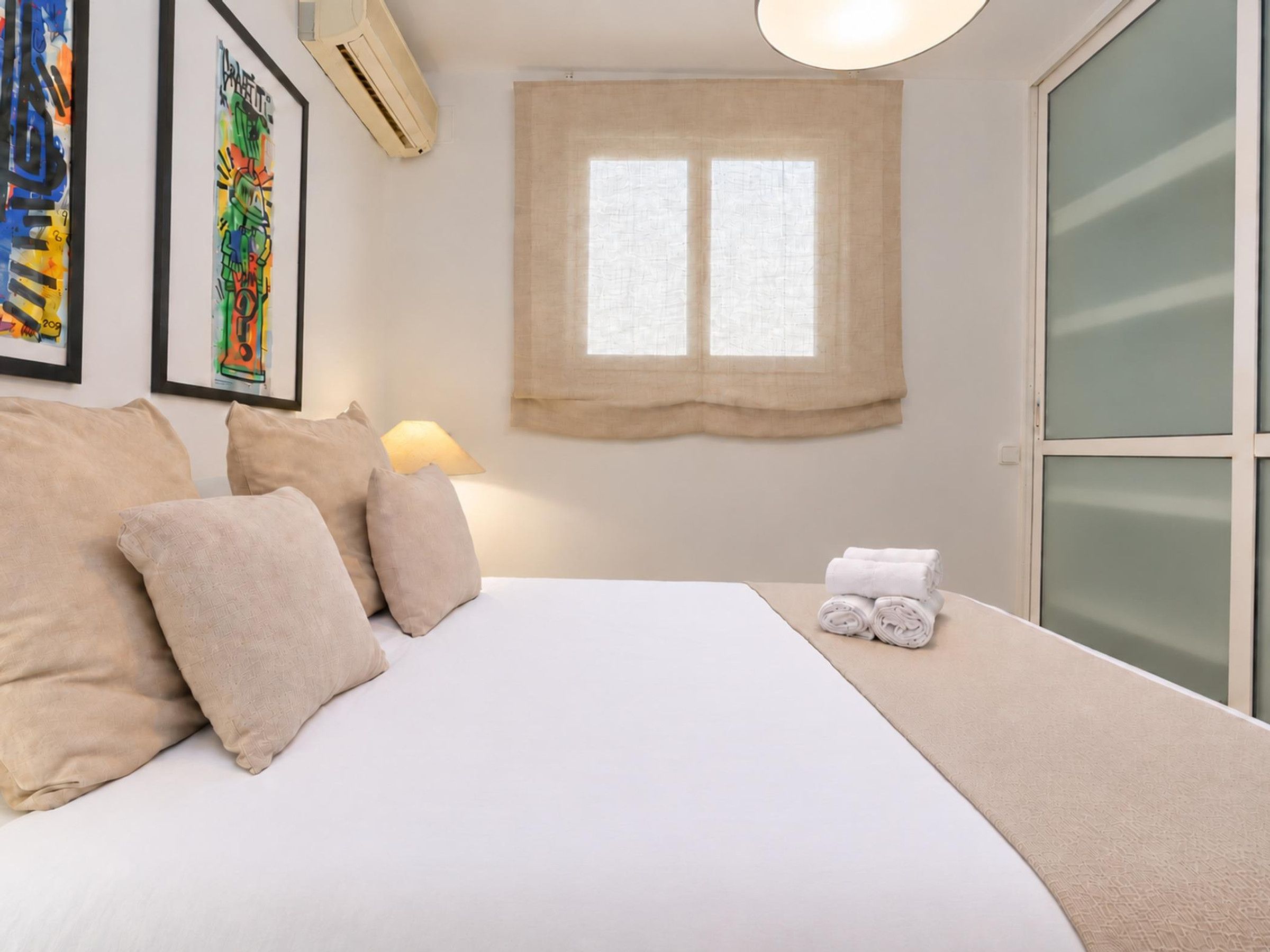 Ein Hotelzimmer mit Bett, Kissen, einem Fenster mit Vorhängen, einer Wand-Klimaanlage und gerahmten Bildern.