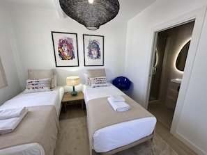 A hotel room with two beds, a small table with a lamp, a blue chair, and a door leading to a bathroom. - Penthouse with panoramic cathedral view - 6 guests (Málaga)