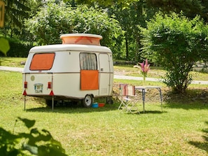 Ein weißer Campervan auf einer Grünfläche, mit orangenen Markisen, einem kleinen Tisch mit Stühlen und einem Blumentopf in der Nähe.