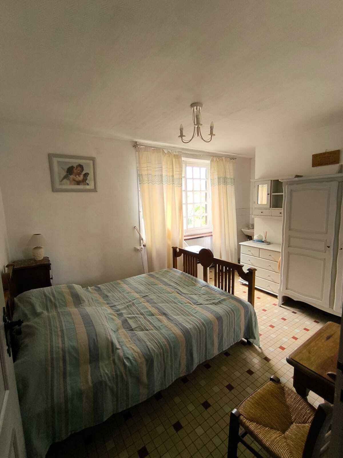 A bedroom with a bed, a chair, a desk, a wardrobe, a window with curtains, a chandelier, and a picture on the wall.