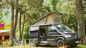 A black van with an open back and a camper trailer attached, parked in a wooded area with a small table and chairs nearby. - Caravan, pets welcome - 4 guests (Séez)